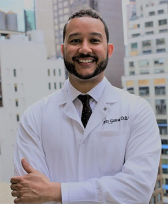 The image is a photograph of a smiling man wearing a white lab coat, standing indoors with a cityscape in the background.