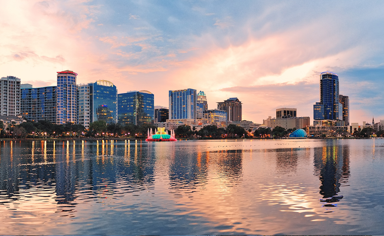 Cityscape with a vibrant skyline and serene lake at sunset, featuring a prominent building and a boat on the water.